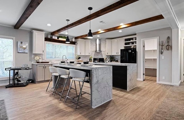 Modern kitchen with wooden floors, marble island, and white cabinetry. Dark wood beams and black pendant lights add contrast, creating a cozy atmosphere.