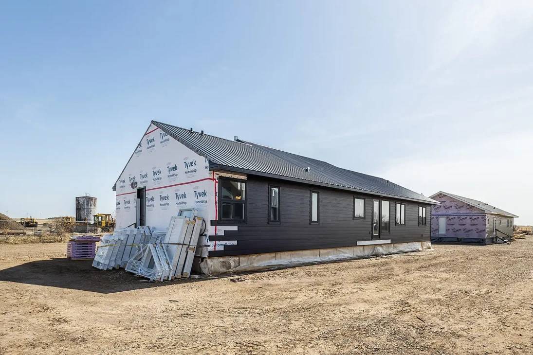 A partially constructed house sits on a barren plot. One side has black siding, while the other is wrapped in Tyvek. Nearby are building materials and another structure.