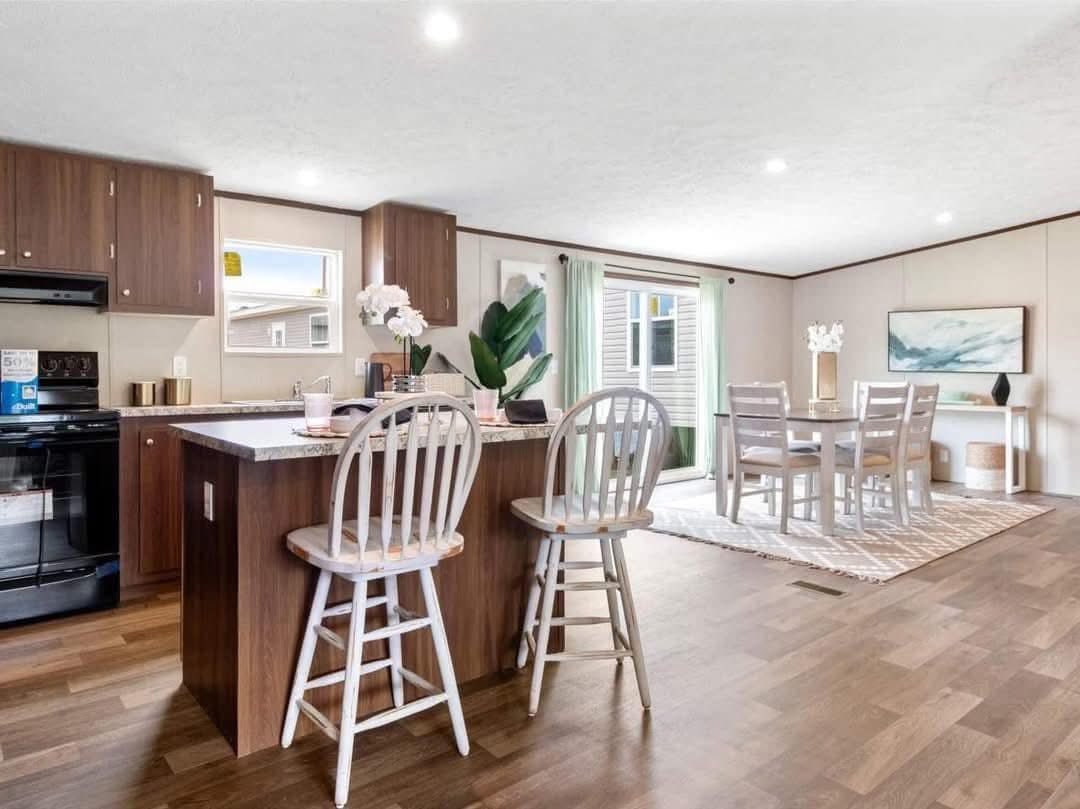 Modern kitchen and dining area with wooden floors. Features an island with barstools, dark cabinets, and a light dining set by a window with green curtains.
