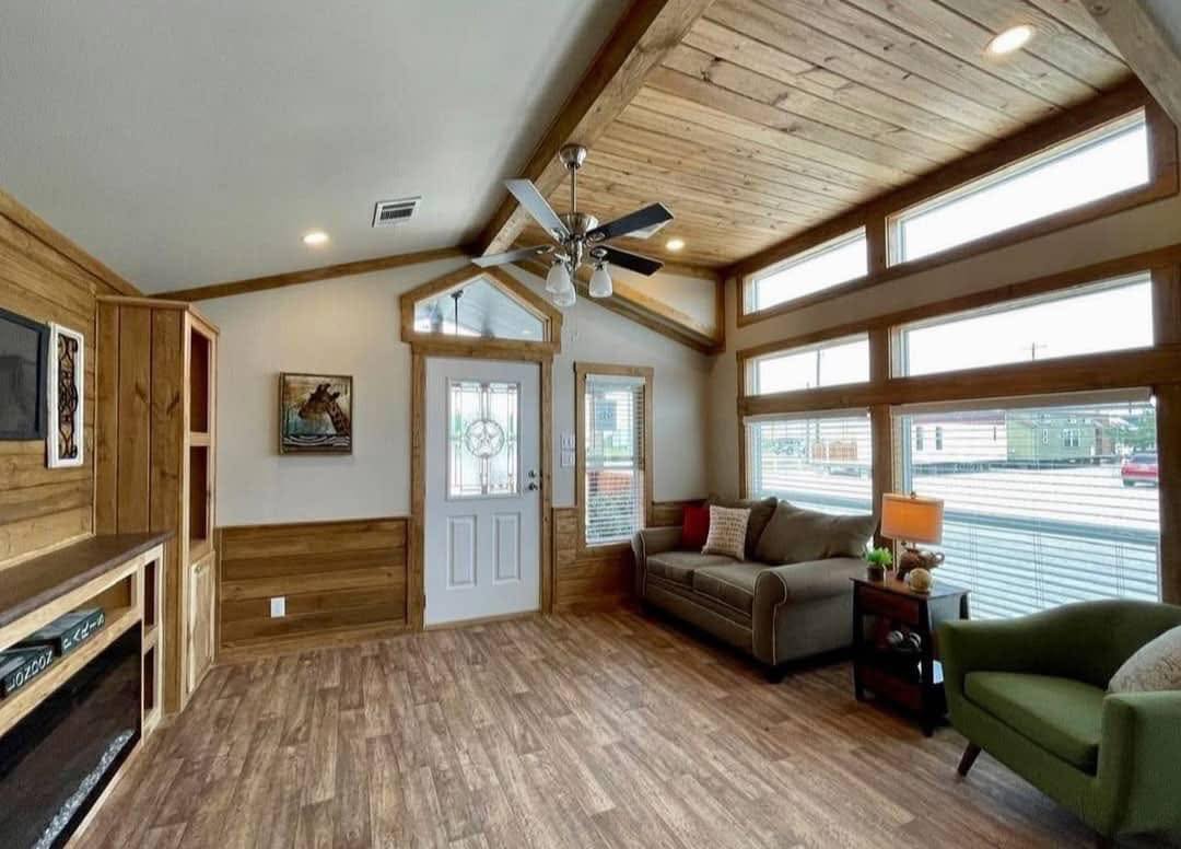 A cozy living room with wooden walls and ceiling, large windows, and a ceiling fan. It features a sofa, green armchair, and warm lighting creating a rustic ambiance.