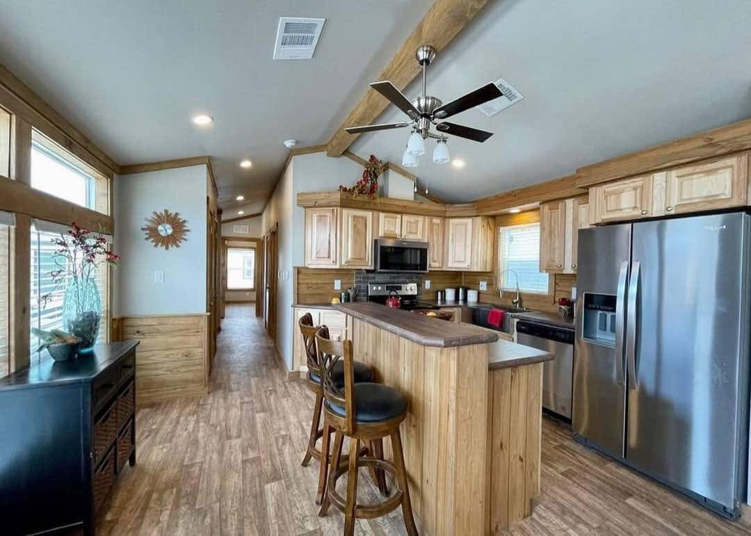 A cozy kitchen with wooden cabinets and accents, bar stools, and stainless steel appliances. Bright ambiance with natural light and a ceiling fan.