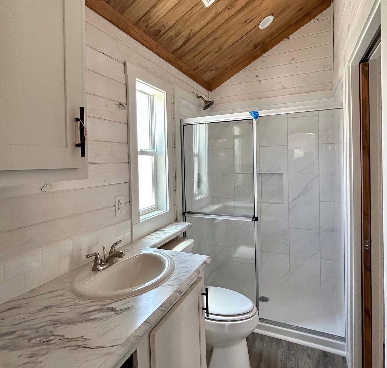 Compact bathroom with a wooden ceiling and white shiplap walls. Features a marble countertop with a sink, a toilet, and a glass-enclosed shower.