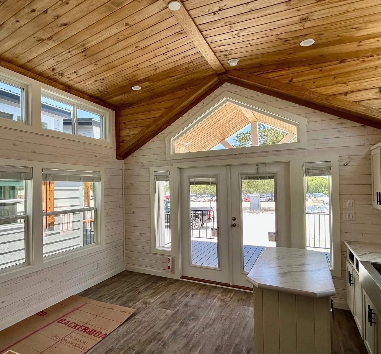 Spacious room with wooden vaulted ceiling, large windows, and glass doors leading outside. Light wood walls and floors create a bright, airy feel.