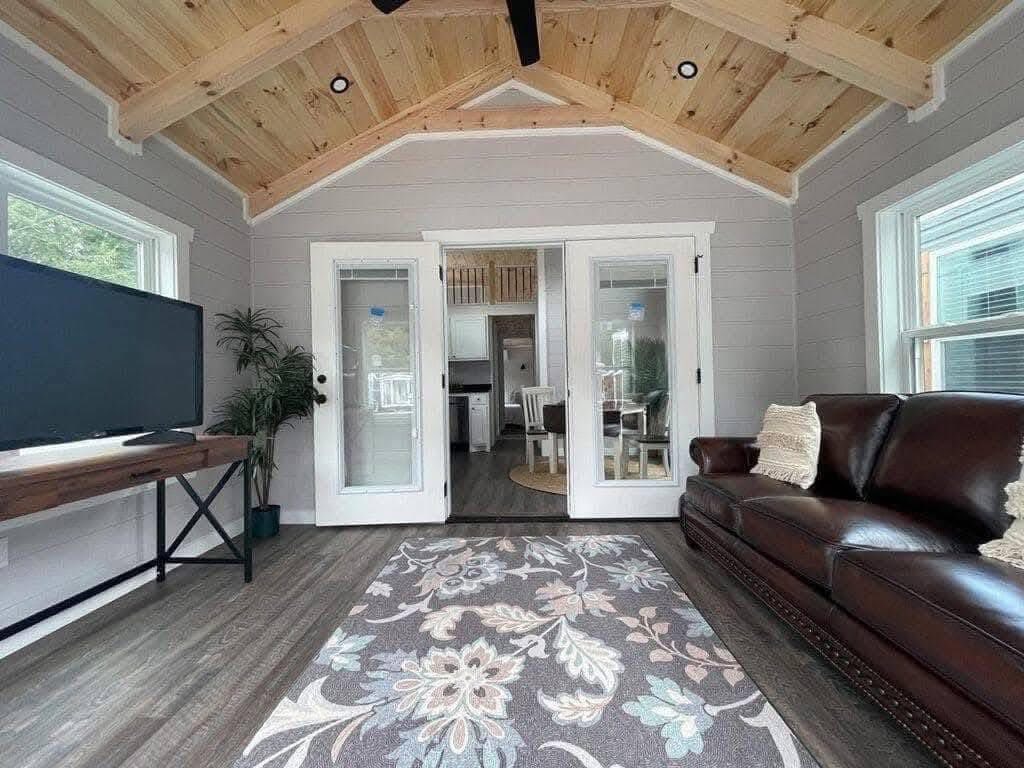 Cozy living room with high wooden ceiling, featuring a brown leather sofa, floral-pattern rug, and TV. Glass doors open to a kitchen area.