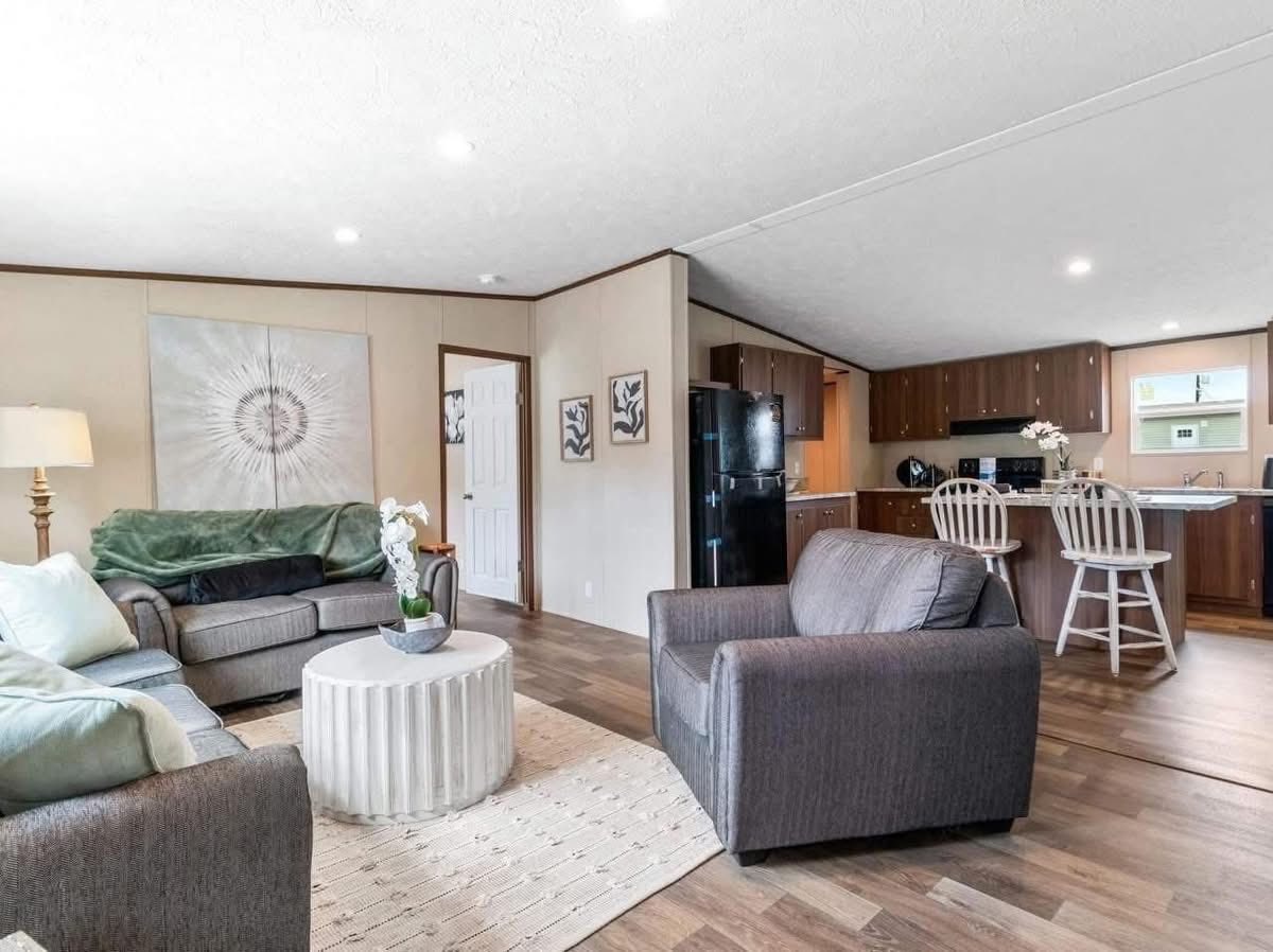 A cozy living room with a gray sofa, green throw, and armchair, surrounded by neutral walls and wooden flooring. A round white coffee table and modern kitchen with bar stools are visible. Soft lighting creates a warm, inviting atmosphere.