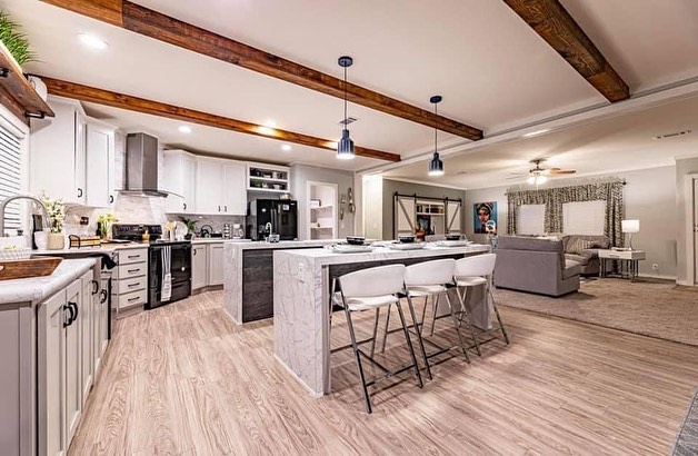 Modern open-plan kitchen and living room with wooden beams. White cabinets, gray island with bar stools, and cozy seating area convey a warm, inviting atmosphere.