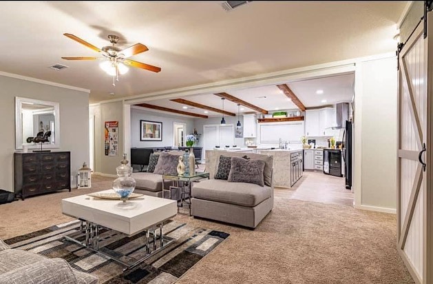 Modern living room with beige carpet and a ceiling fan. Cozy gray sectional sofas face a sleek white coffee table. In the background, a spacious kitchen with white cabinets and wooden ceiling beams adds a warm touch. A barn door is visible on the right, contributing to the rustic, inviting atmosphere.