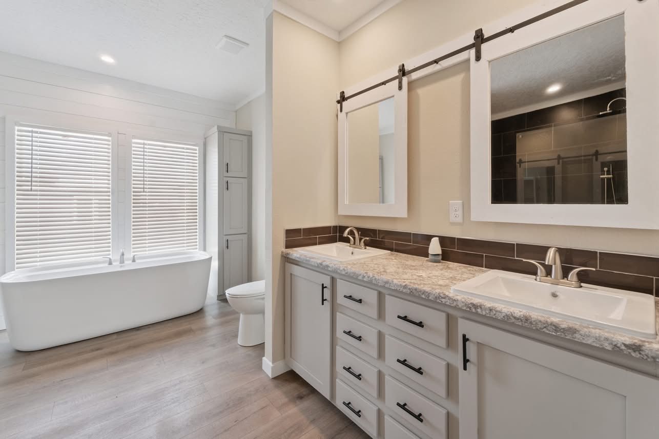 Spacious bathroom with a freestanding white tub, large windows with blinds, a double vanity with two sinks and mirrors, and light wood flooring.