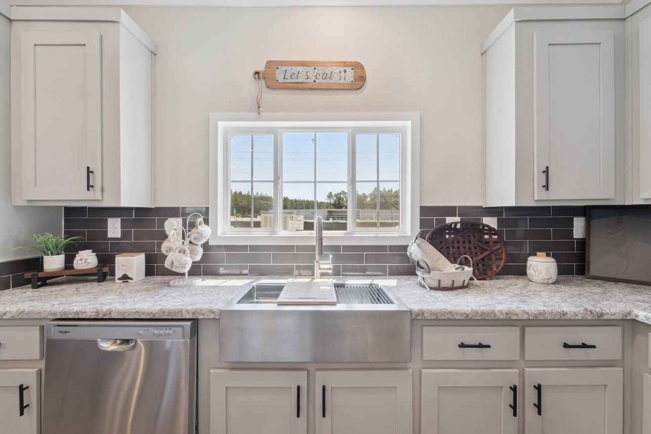 Bright kitchen with white cabinets, marble countertops, and stainless steel sink. Subway tile backsplash. Window offers a view outside. Cozy, inviting mood.