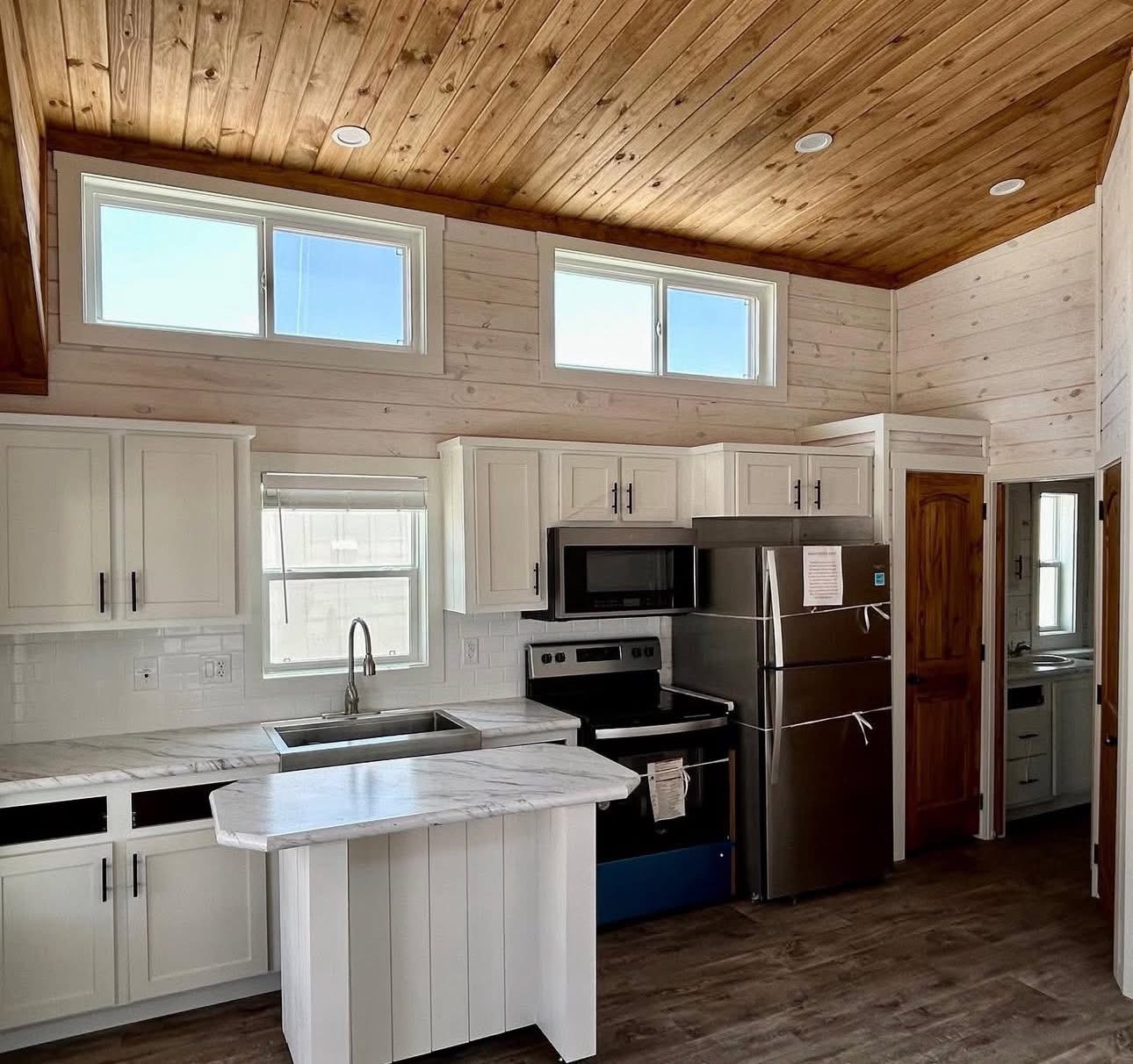 Modern kitchen with wood-paneled ceiling, white cabinets, and stainless steel appliances. Large windows provide ample natural light. Elegant and cozy ambiance.