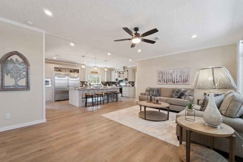 Spacious living room and kitchen with light wood flooring. Cozy beige sofa, modern decor, a ceiling fan, and pendant lights create a warm, inviting atmosphere.