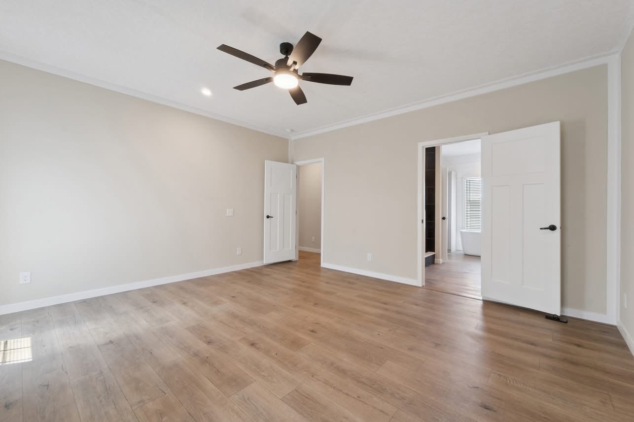 A spacious, empty room with light beige walls, wood flooring, and a ceiling fan. Two open doors reveal a hallway and a bathroom, creating a calm atmosphere.