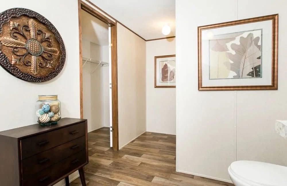 A minimalist room features a wooden dresser with decorative shells, framed leaf art, and a carved wall piece. The open door leads to a closet.
