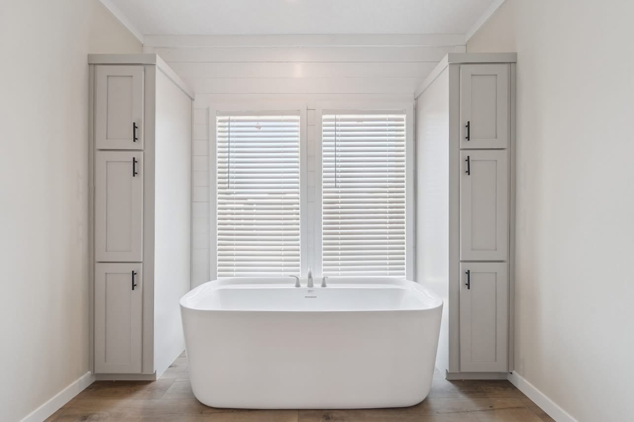 A modern bathroom features a sleek white freestanding tub between tall gray cabinets. The room is brightened by sunlight filtering through white blinds, evoking tranquility.