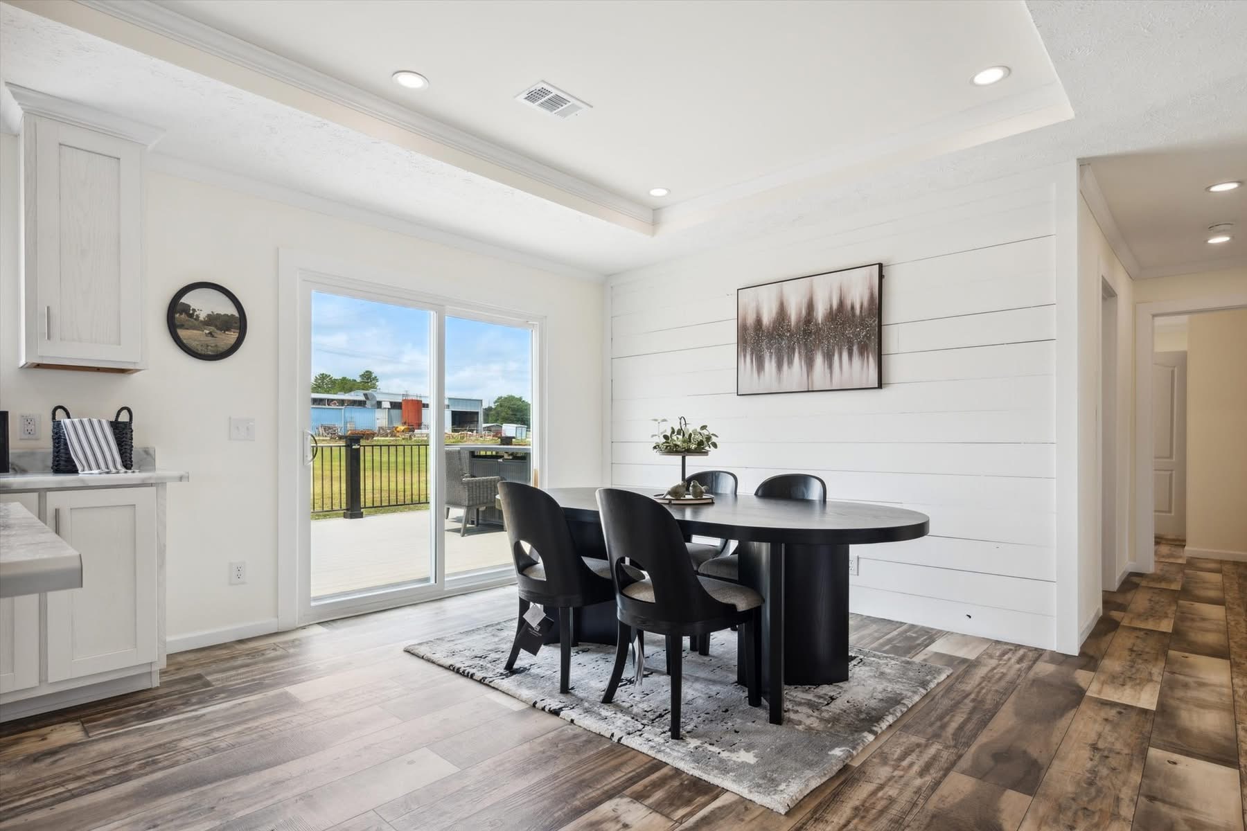 Modern dining area with a round black table, four matching chairs on a patterned rug, white shiplap walls, large glass doors, and minimalistic decor.