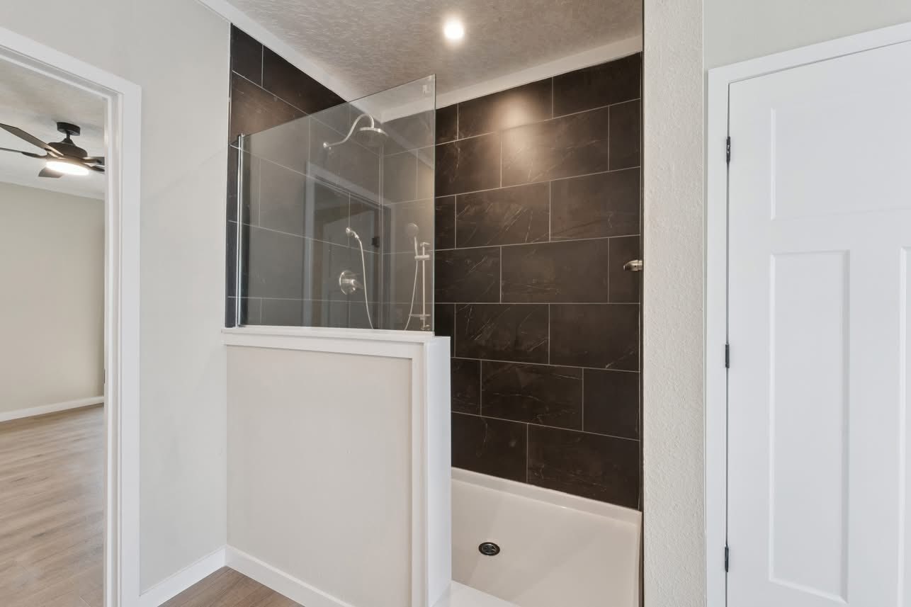 Modern bathroom with a glass-enclosed shower featuring dark tiles and a chrome showerhead. Adjacent is an open doorway leading to a room with wood flooring.