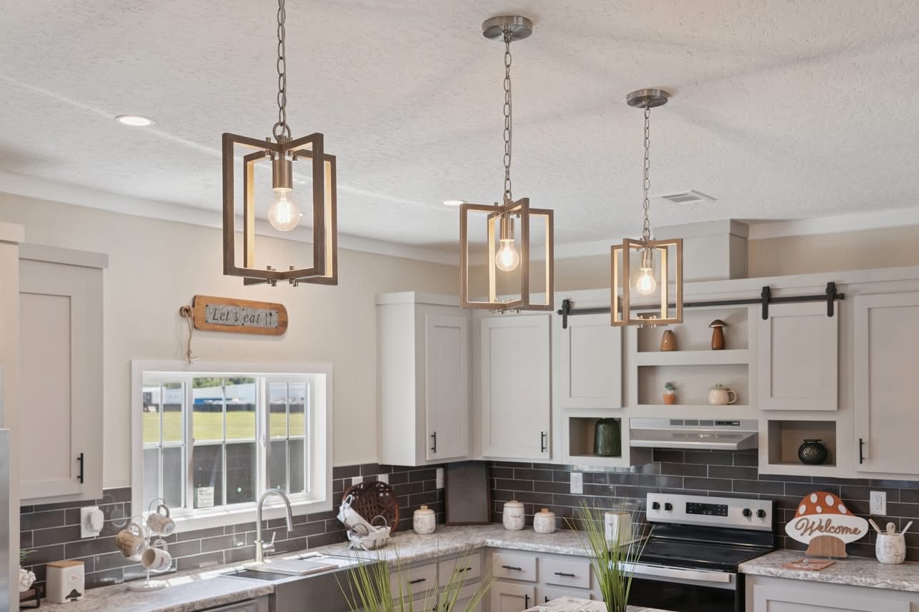 Modern kitchen with white cabinets, gray tile backsplash, and three pendant lights. Cozy decor includes welcome sign and small plants on the countertops.