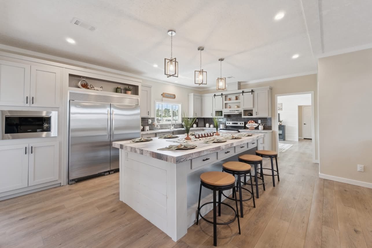 Modern open-plan living room and kitchen with neutral tones. Features a cozy sofa, large rug, round coffee table, island with stools, and hanging lights.