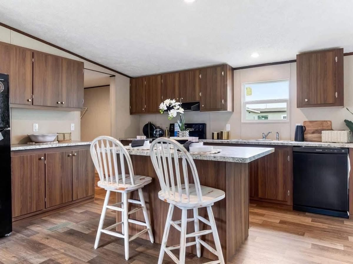 A cozy kitchen with brown cabinets and an L-shaped granite countertop. Two white chairs face an island adorned with flowers, creating a welcoming atmosphere.