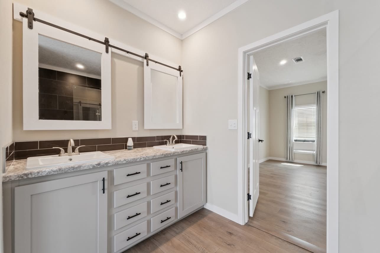 Modern bathroom with a double-sink vanity, gray cabinets, and wall-mounted sliding mirrors. An open door leads to a bright room with wooden flooring.