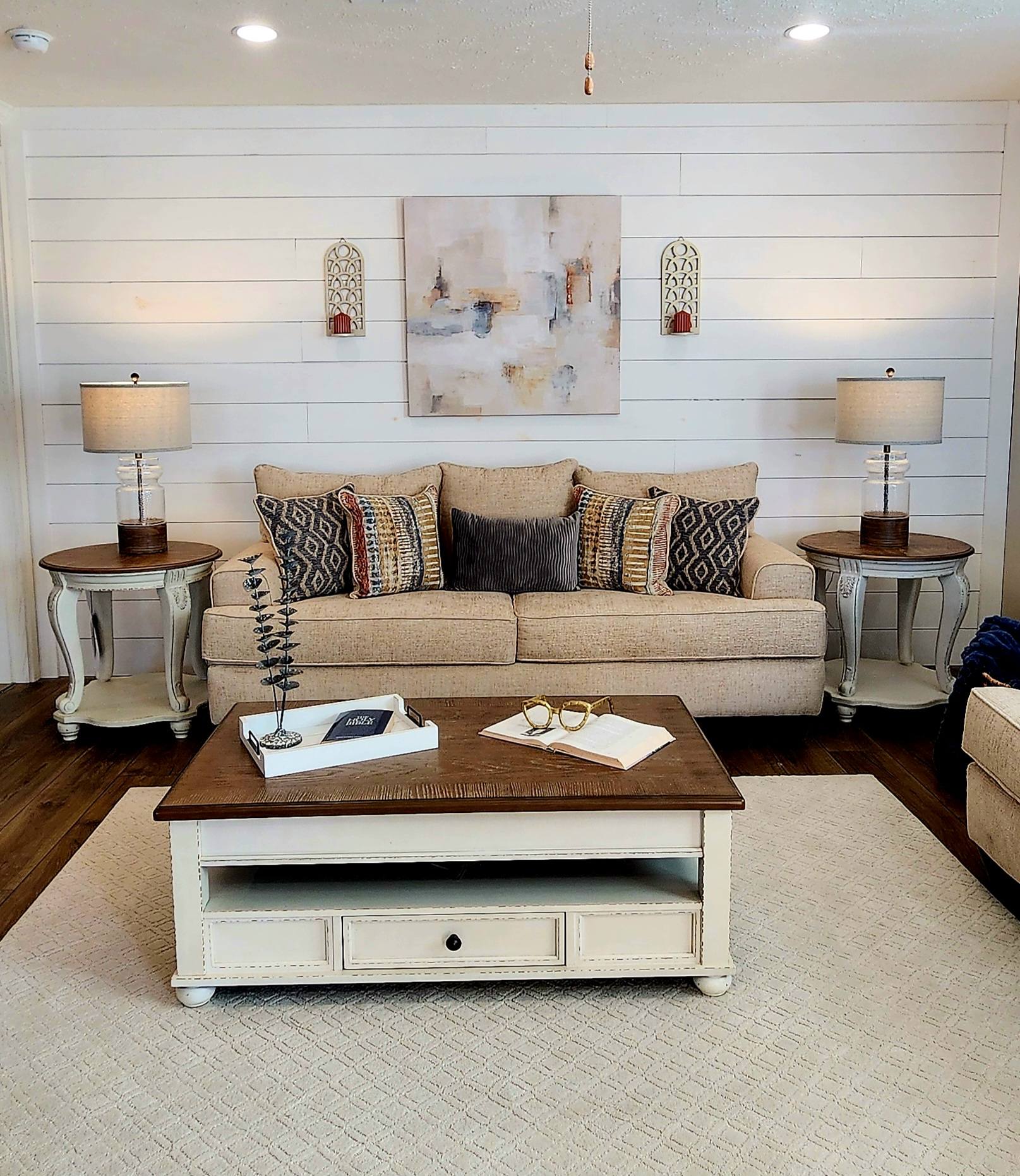 Cozy living room with a beige sofa, colorful patterned pillows, and wooden side tables. A soft rug and abstract art add warmth and elegance.