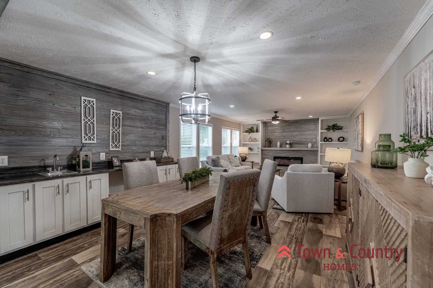 Stylish open-plan room featuring a rustic wooden dining table with upholstered chairs, modern light fixtures, and cozy gray sofas near a fireplace. The ambiance is warm and inviting.