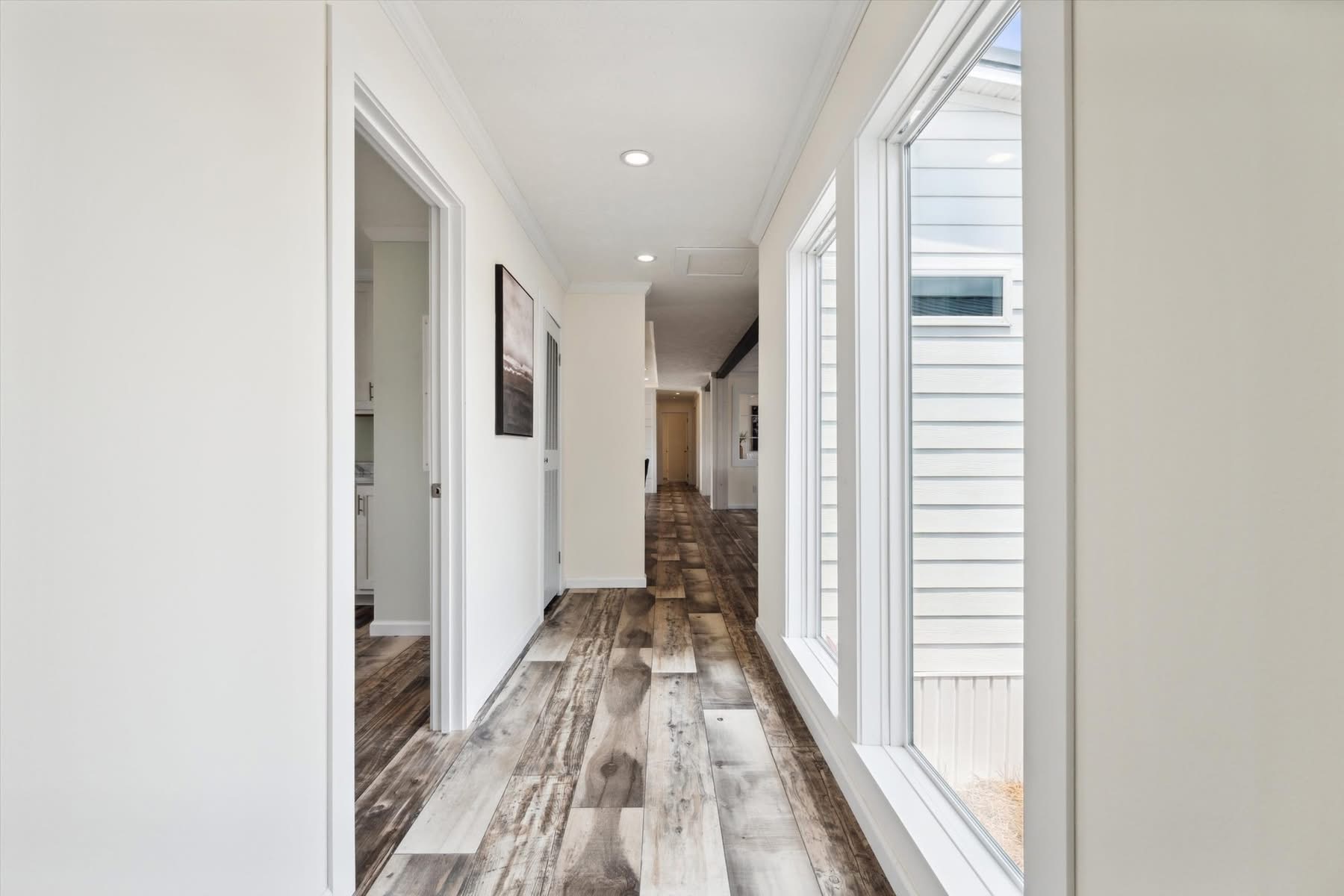 Bright hallway with wood flooring, white walls, large windows on the right letting in sunlight, and doorways to the left. Minimalist and airy.