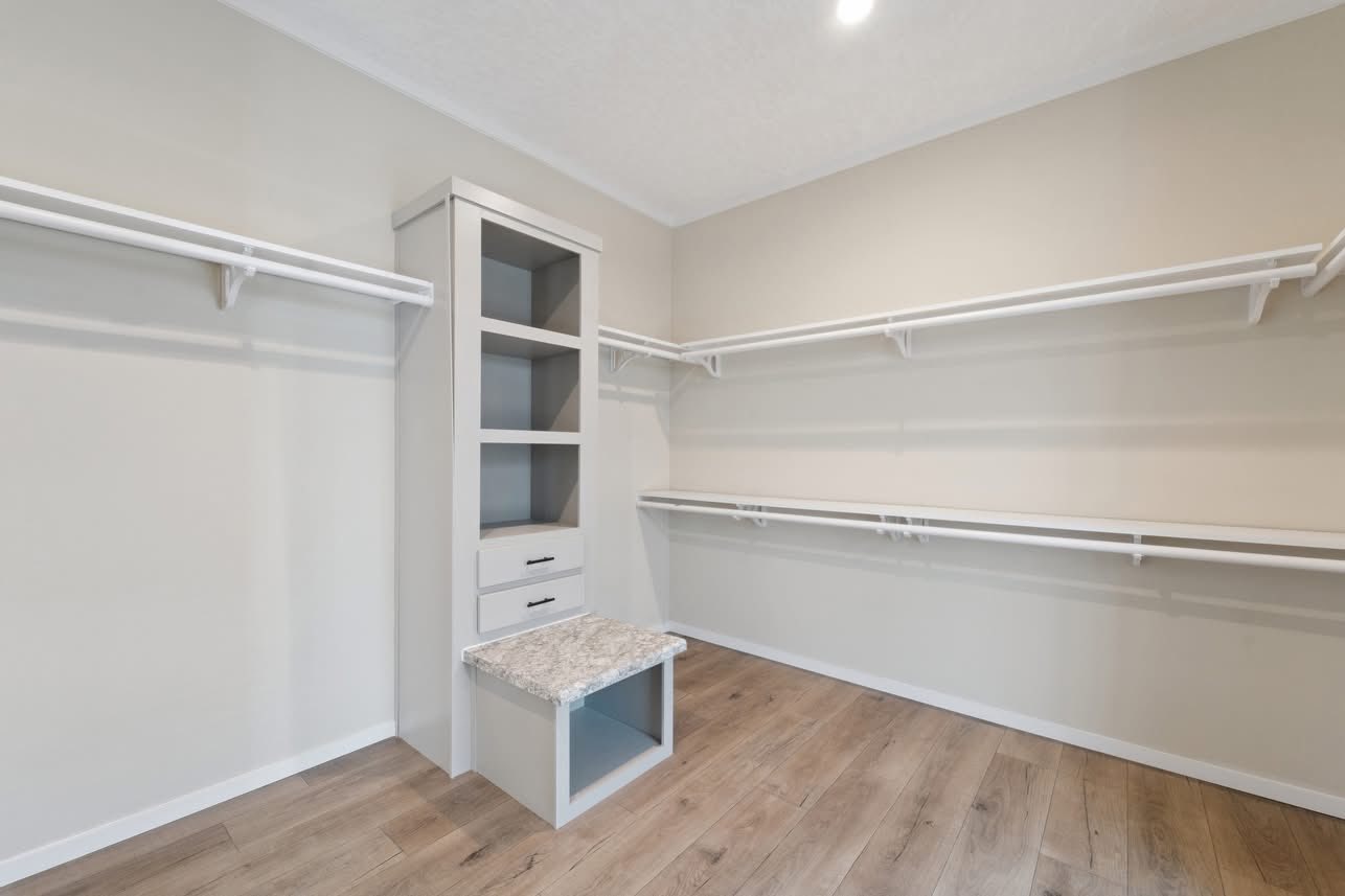 Spacious walk-in closet with light wood flooring, neutral walls, and white shelves. Central unit with open shelves, drawers, and a gray cushioned bench.