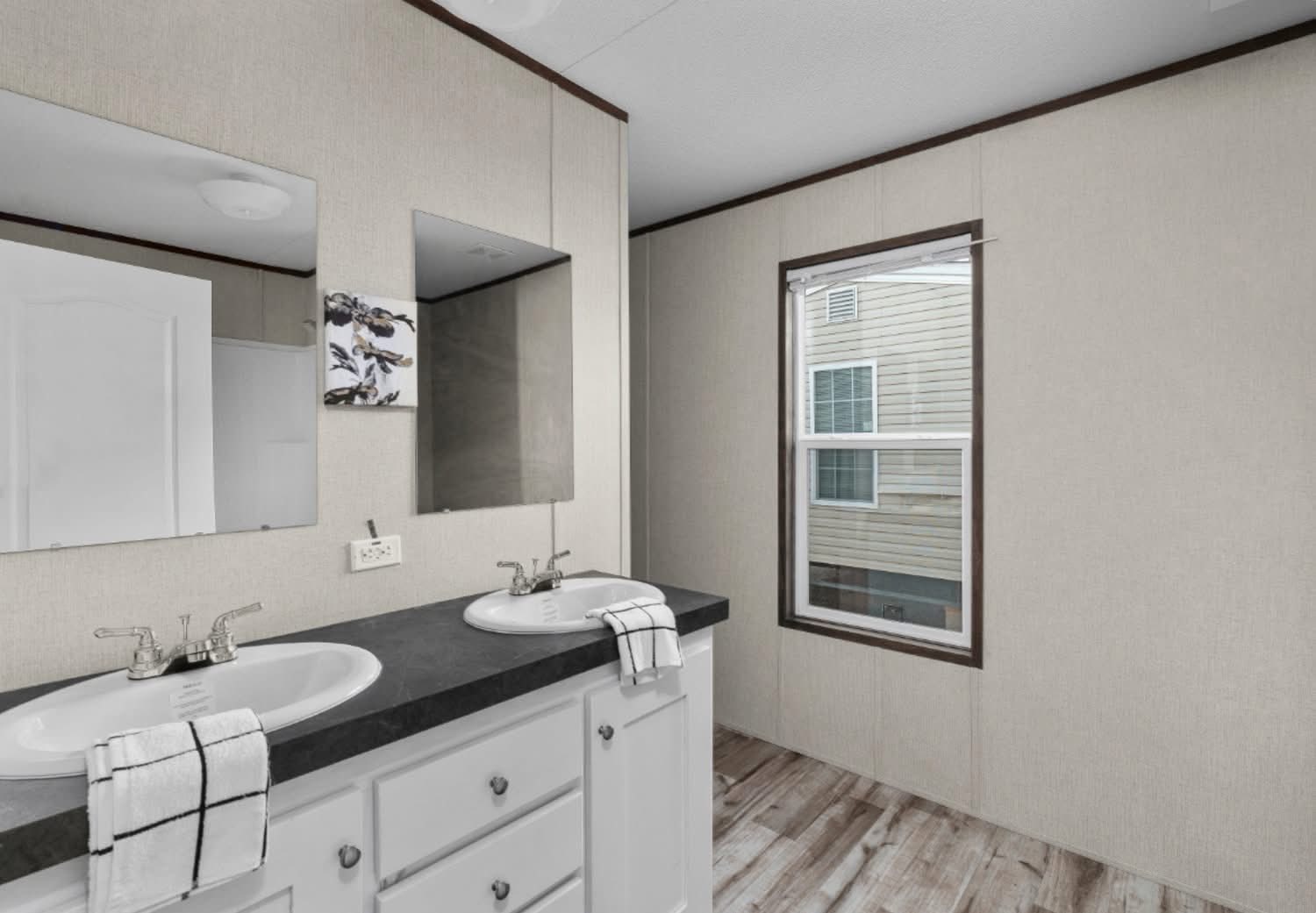 Modern bathroom with double vanity, black countertop, and two sinks. Neutral walls, wood floor, large window, and a small wall painting. Clean and minimalistic.