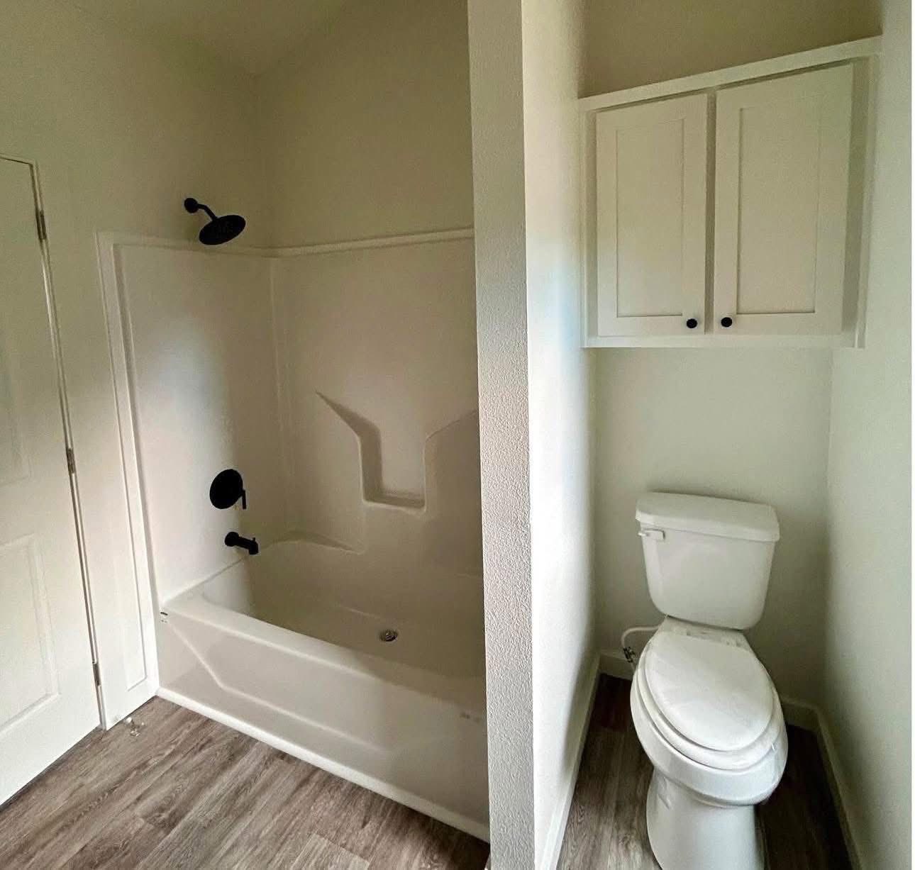 A small bathroom with a white bathtub and black fixtures on the left, a toilet on the right, and white cabinets above it. The floor has wood-like tiles.