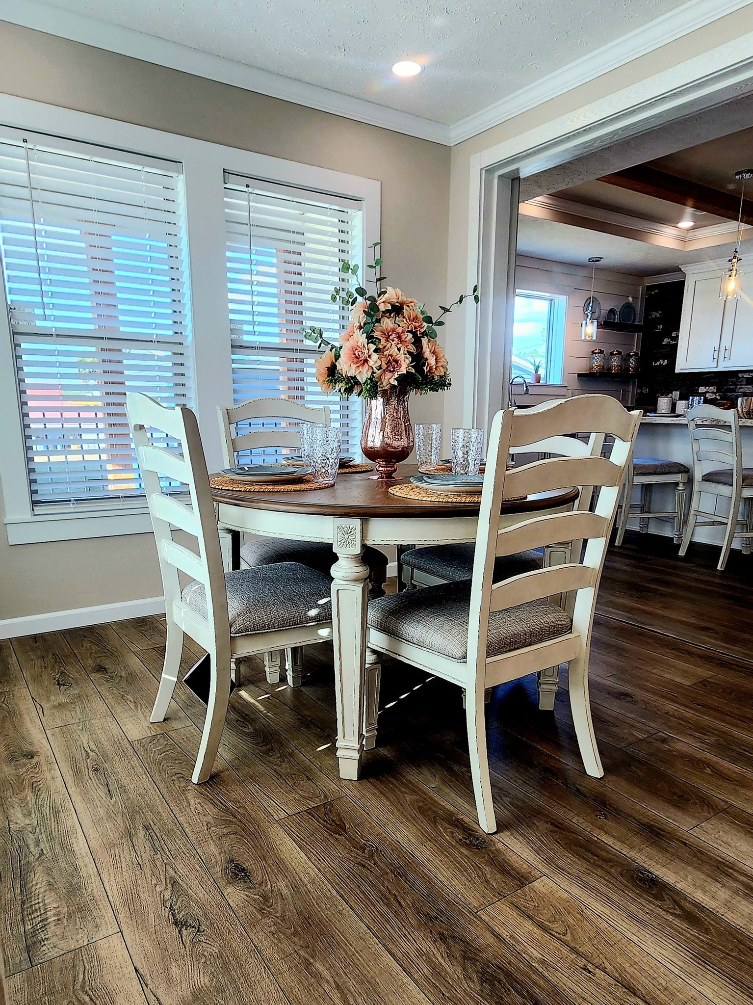 Cozy dining area with a round wooden table, four cushioned chairs, a vase of pink flowers, surrounded by bright windows and warm wooden flooring.