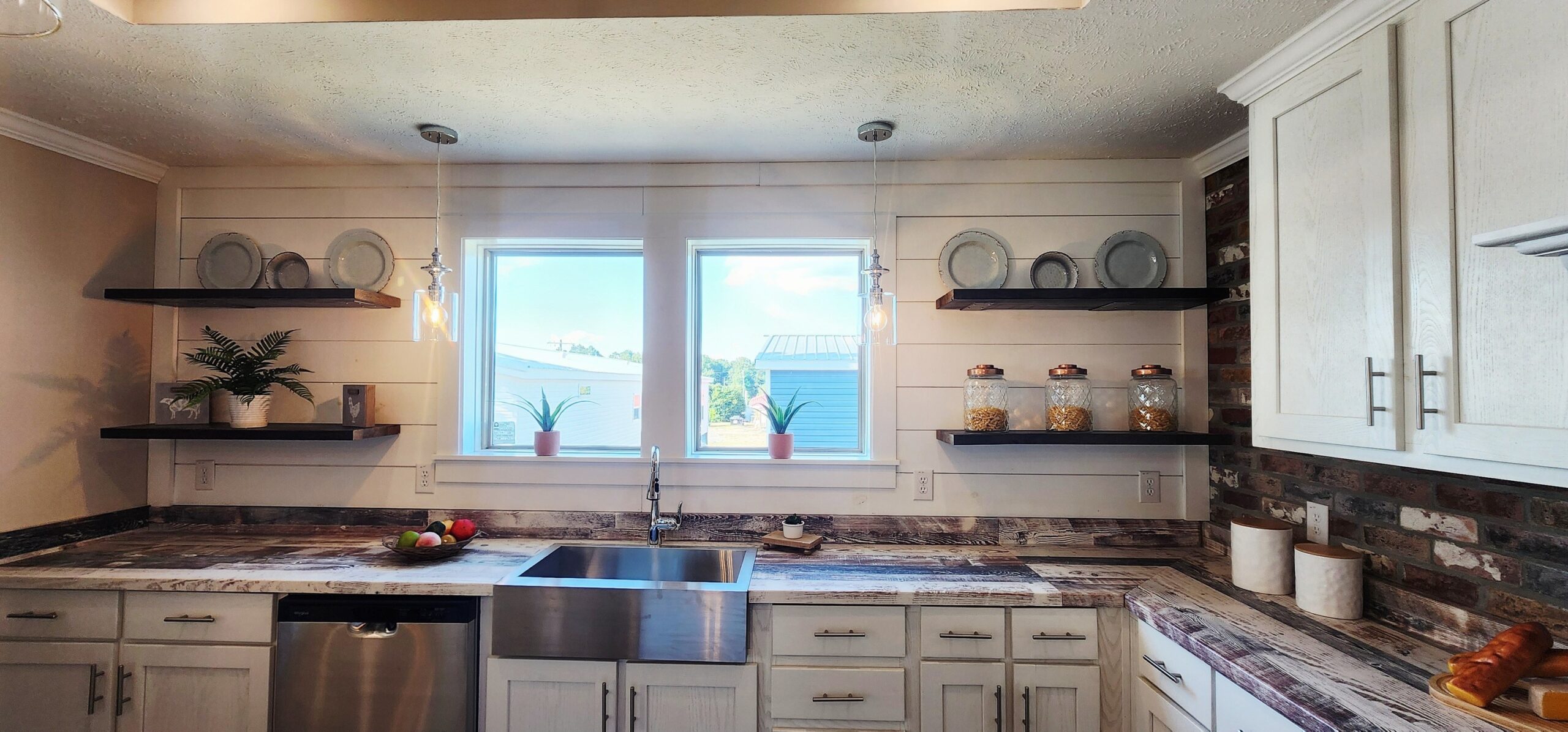 Modern kitchen with white cabinets, rustic shelves, and a farmhouse sink. Sunlight streams through two windows, creating a bright, cozy atmosphere.