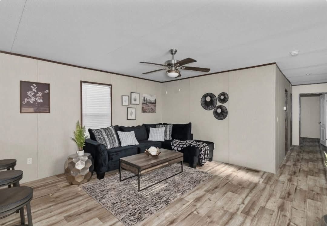 Modern living room with a dark blue sectional sofa, patterned pillows, and a coffee table on a textured rug. Wall art and a ceiling fan add decor. Cozy atmosphere.