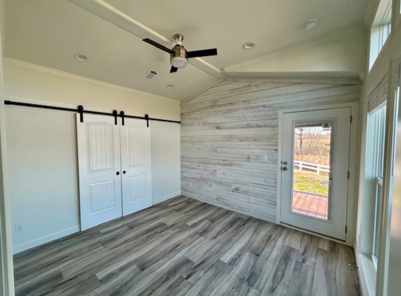 A bright, empty room with wood-look flooring, a ceiling fan, two sliding barn doors, and a rustic wood accent wall. A glass door opens to a sunny yard.