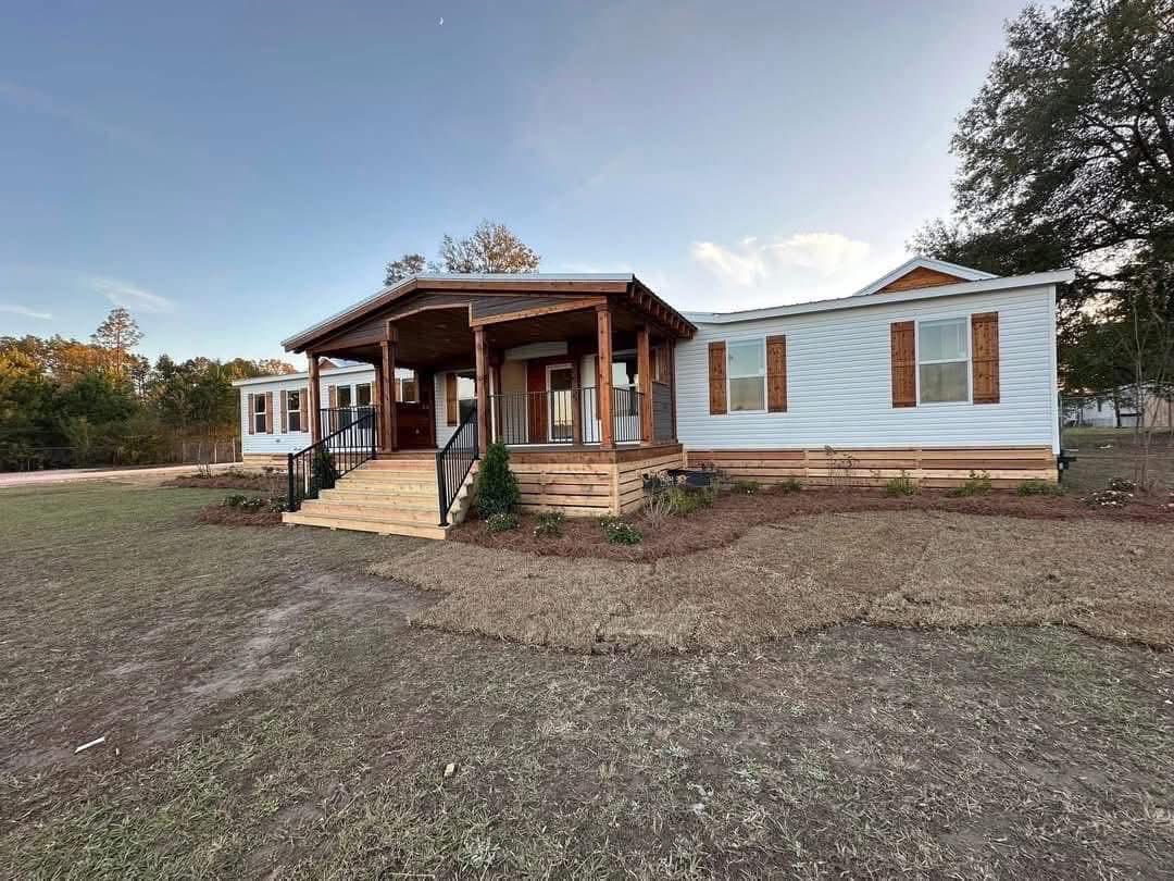 Single-story modular home with white siding and wooden shutters, featuring a welcoming porch with wooden pillars. Set on a spacious lawn under a clear sky.