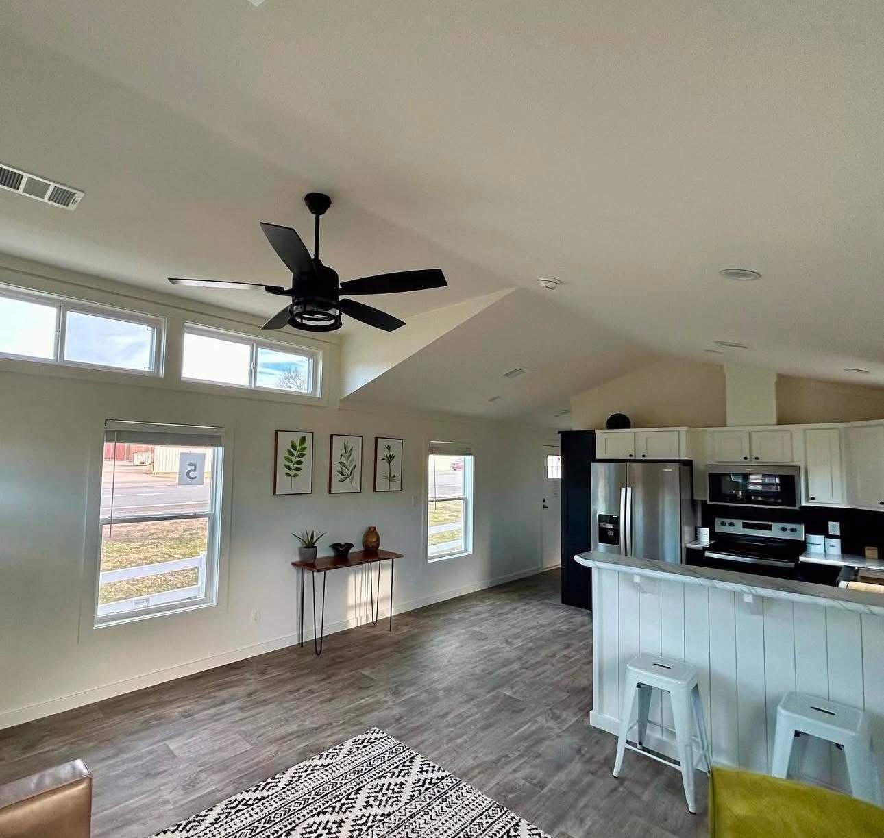 Modern living room and kitchen with high ceilings and natural light. Features a black ceiling fan, botanical art, stainless steel appliances, and wood flooring.