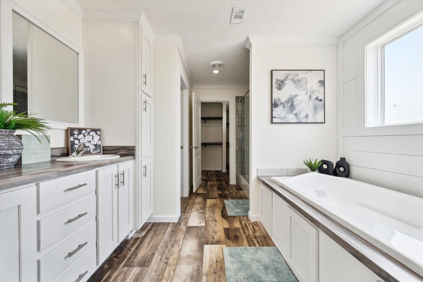 Spacious modern bathroom with wooden floors, white cabinets, and a large bathtub. A window provides natural light; minimalist decor adds elegance.
