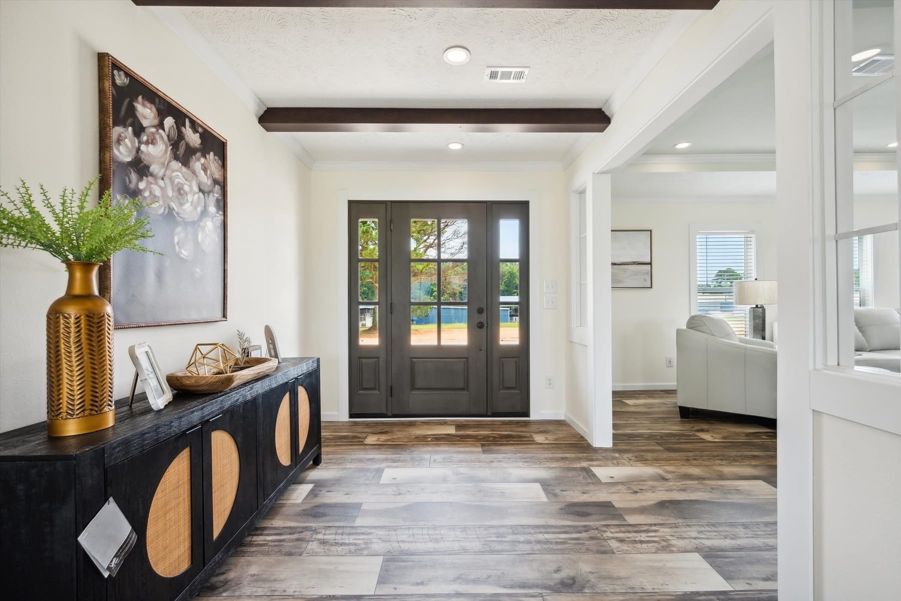 Bright entryway with dark wood door and sidelights. Modern black console table with decor sits beneath floral art. Cozy adjacent living room visible.