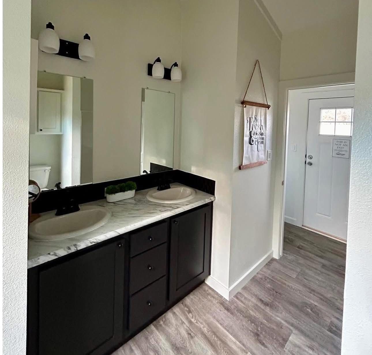 Modern bathroom with dual sinks on a marble countertop, black cabinets, and round mirrors. Bright lighting and wooden flooring create a fresh ambiance.
