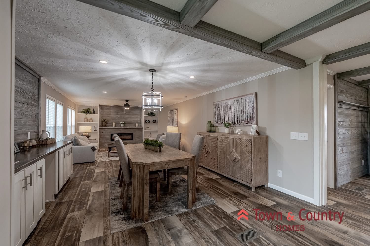 Spacious dining and living area with rustic wood floors and ceiling beams. Features a long wooden table, modern chandelier, and cozy seating by a fireplace.