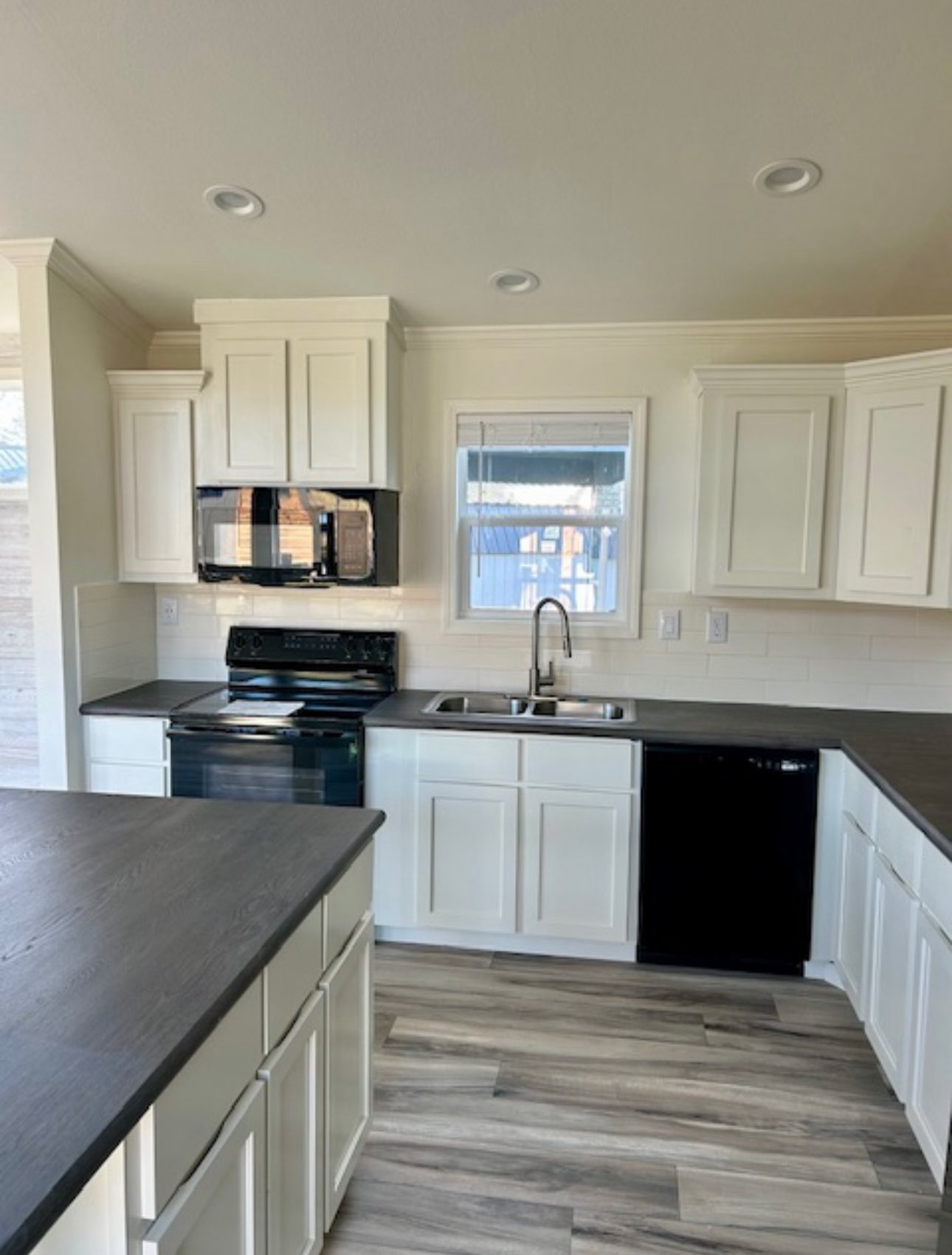 Modern kitchen with white cabinets, black appliances, and a dark countertop. Features a central window, wood-style flooring, and a clean, bright atmosphere.