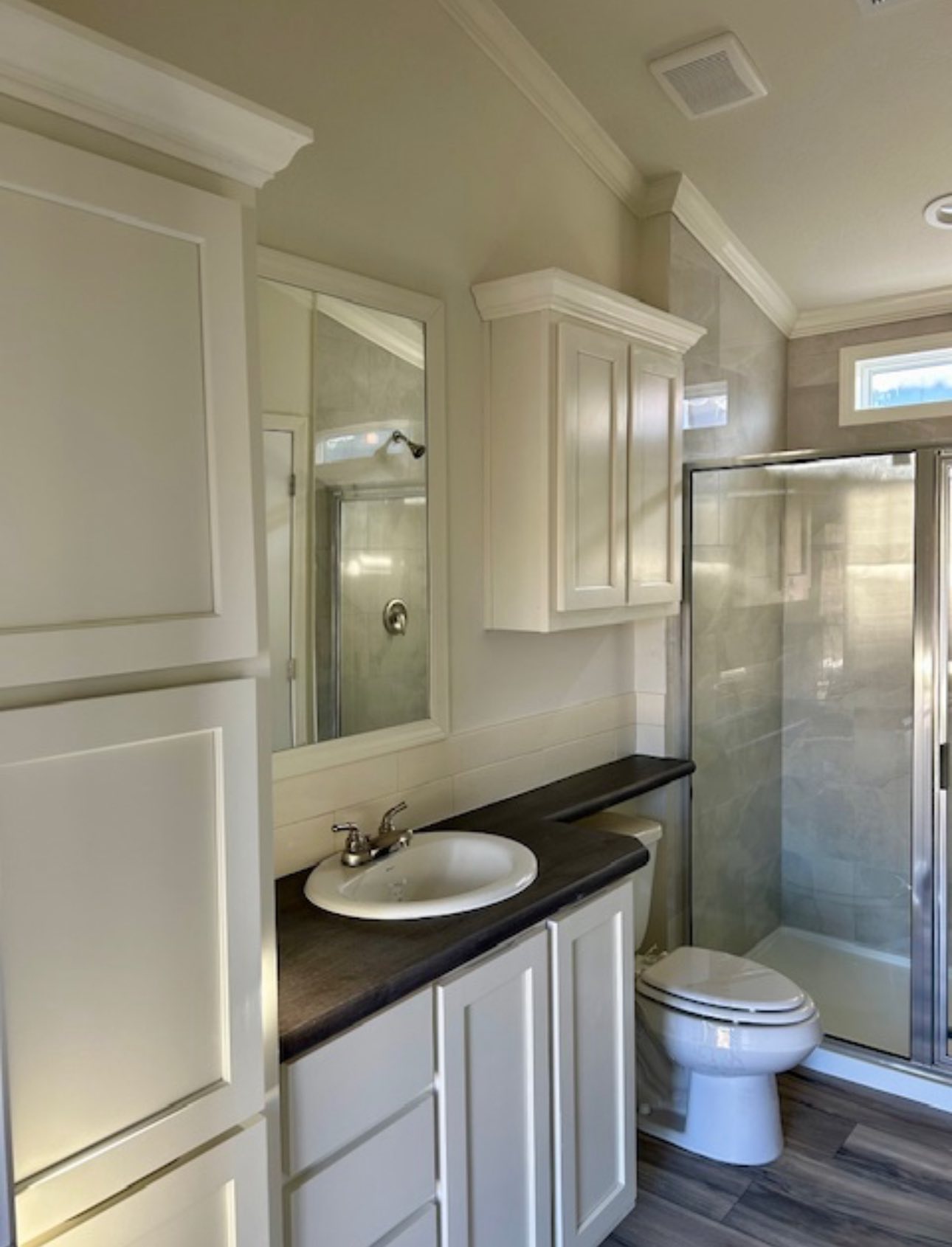 A bright bathroom with beige cabinets, a dark countertop, a white sink, and a glass shower. The space is modern and orderly, conveying a clean ambiance.