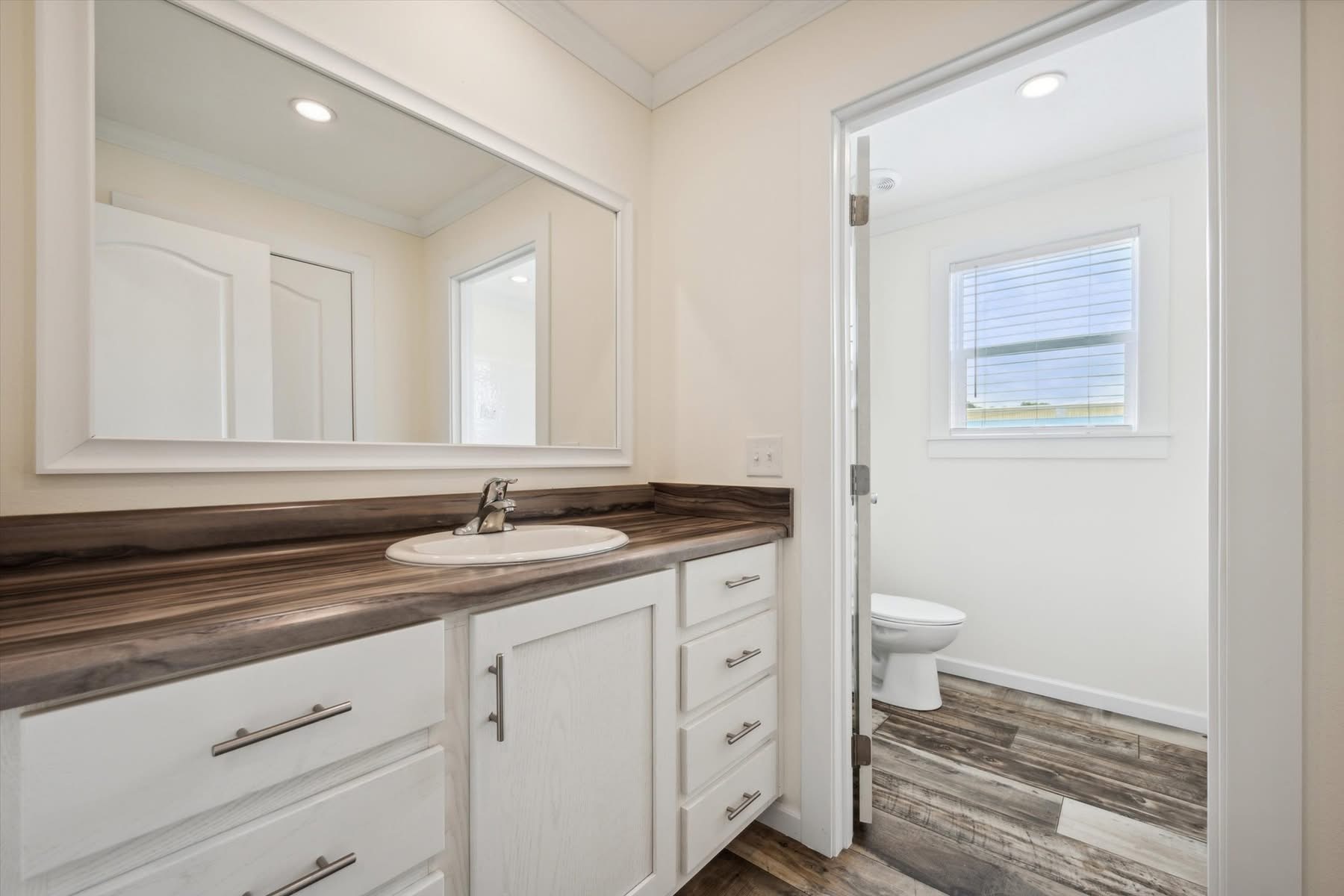 Modern bathroom with a white vanity, dark wooden countertop, and sink. A large mirror above and door leading to a room with a window and wooden floor. Warm, clean ambiance.