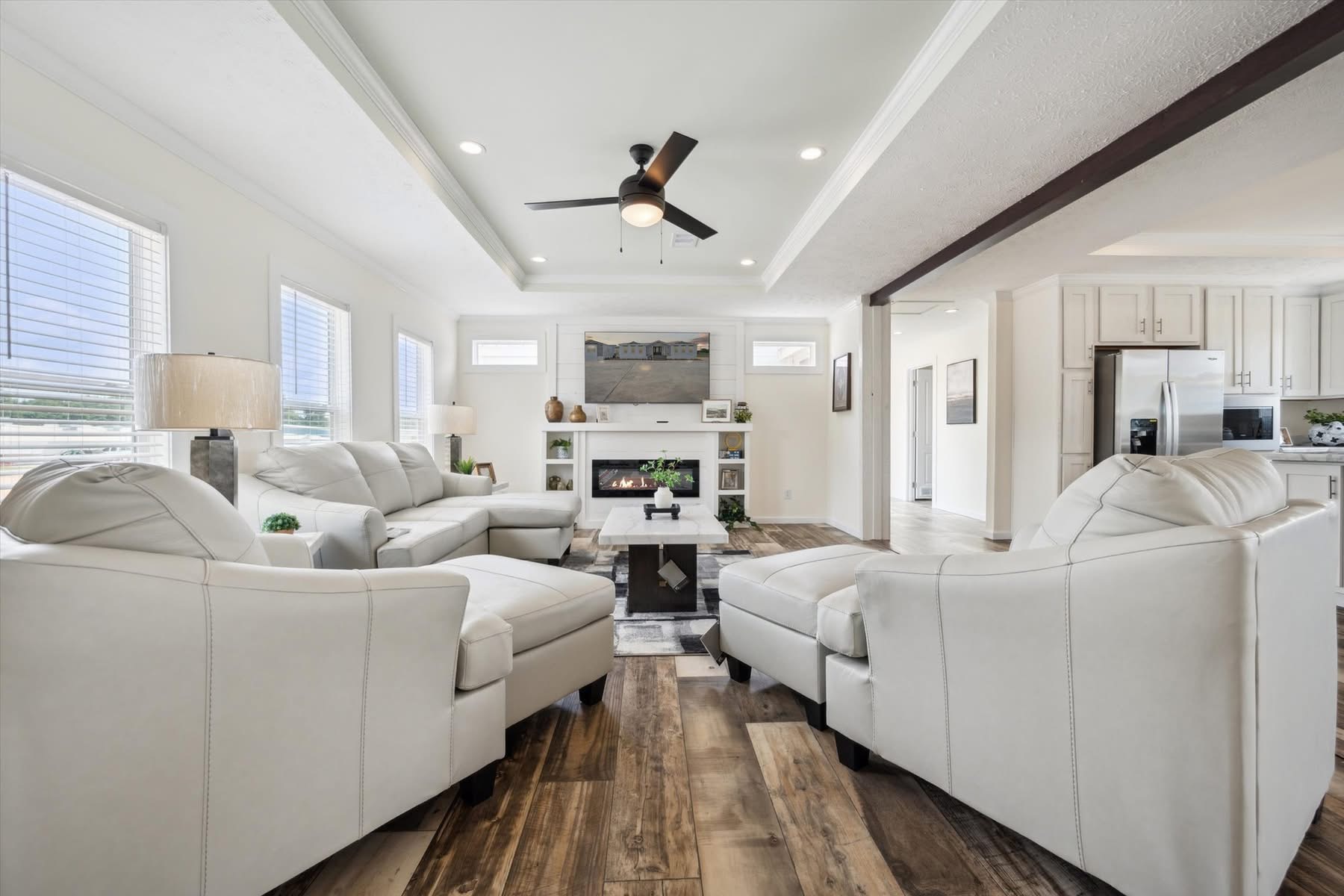 Stylish living room with white leather sofas, wooden flooring, a ceiling fan, and a cozy fireplace. Bright ambiance with natural light.