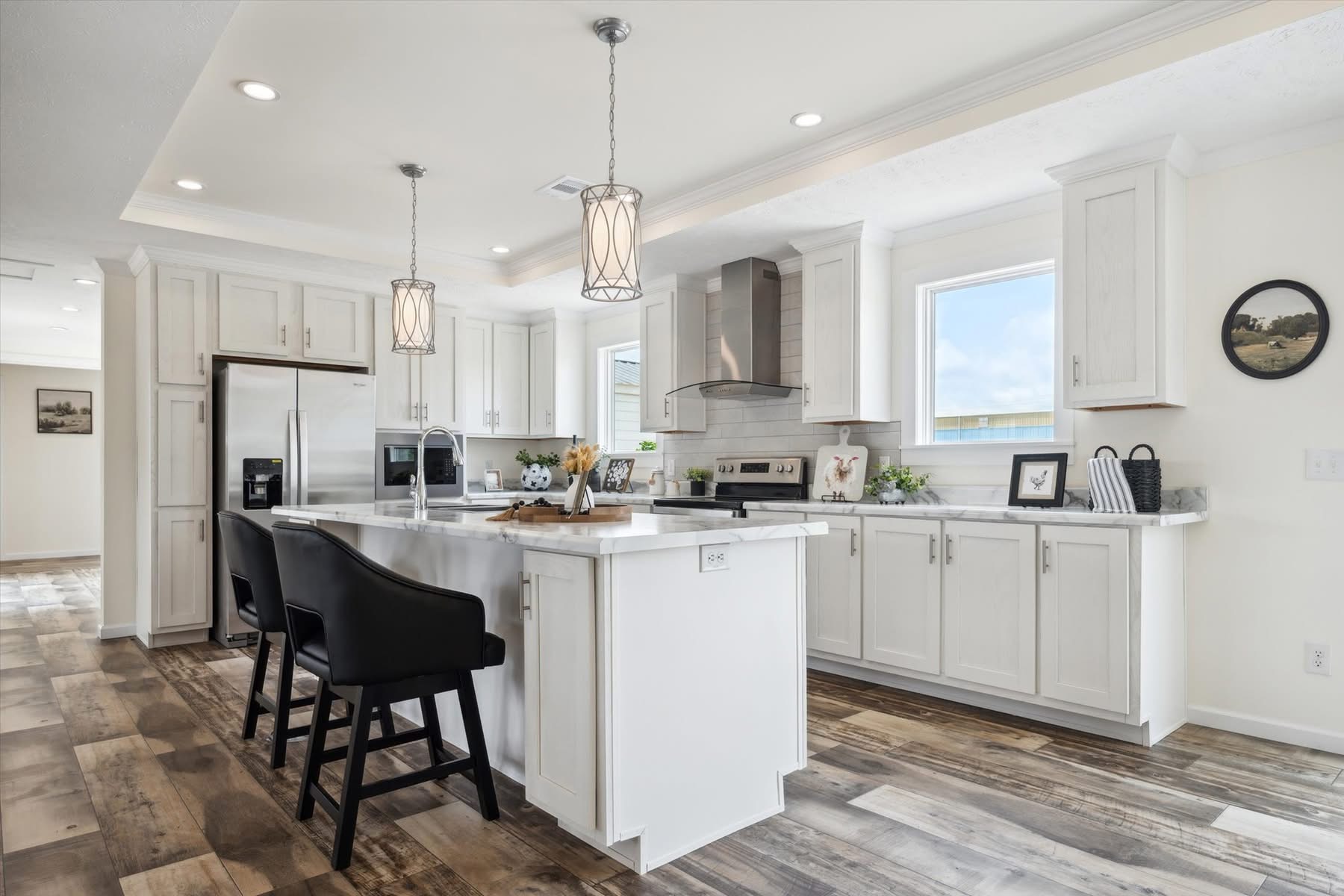 A modern kitchen with white cabinets, wood flooring, and an island with two black chairs. Two pendant lights hang above the island. Natural light streams through a large window, creating an inviting atmosphere.