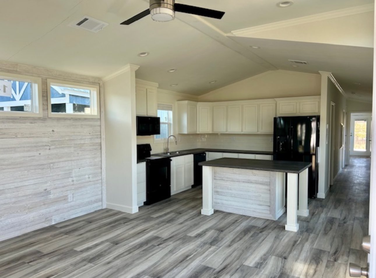 A modern kitchen with white cabinets, black appliances, and a central island, featuring a light wood floor and ceiling fan, creating a spacious, airy feel.