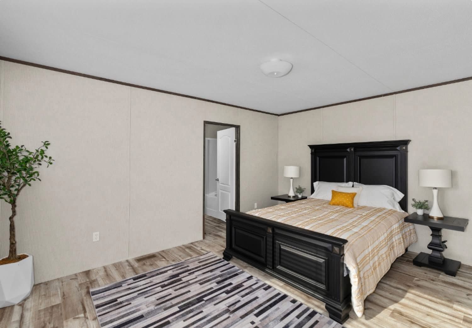 A spacious bedroom with a large black bed featuring plaid bedding and a yellow cushion. Two bedside tables with lamps flank the bed. A striped rug and a potted plant add warmth.
