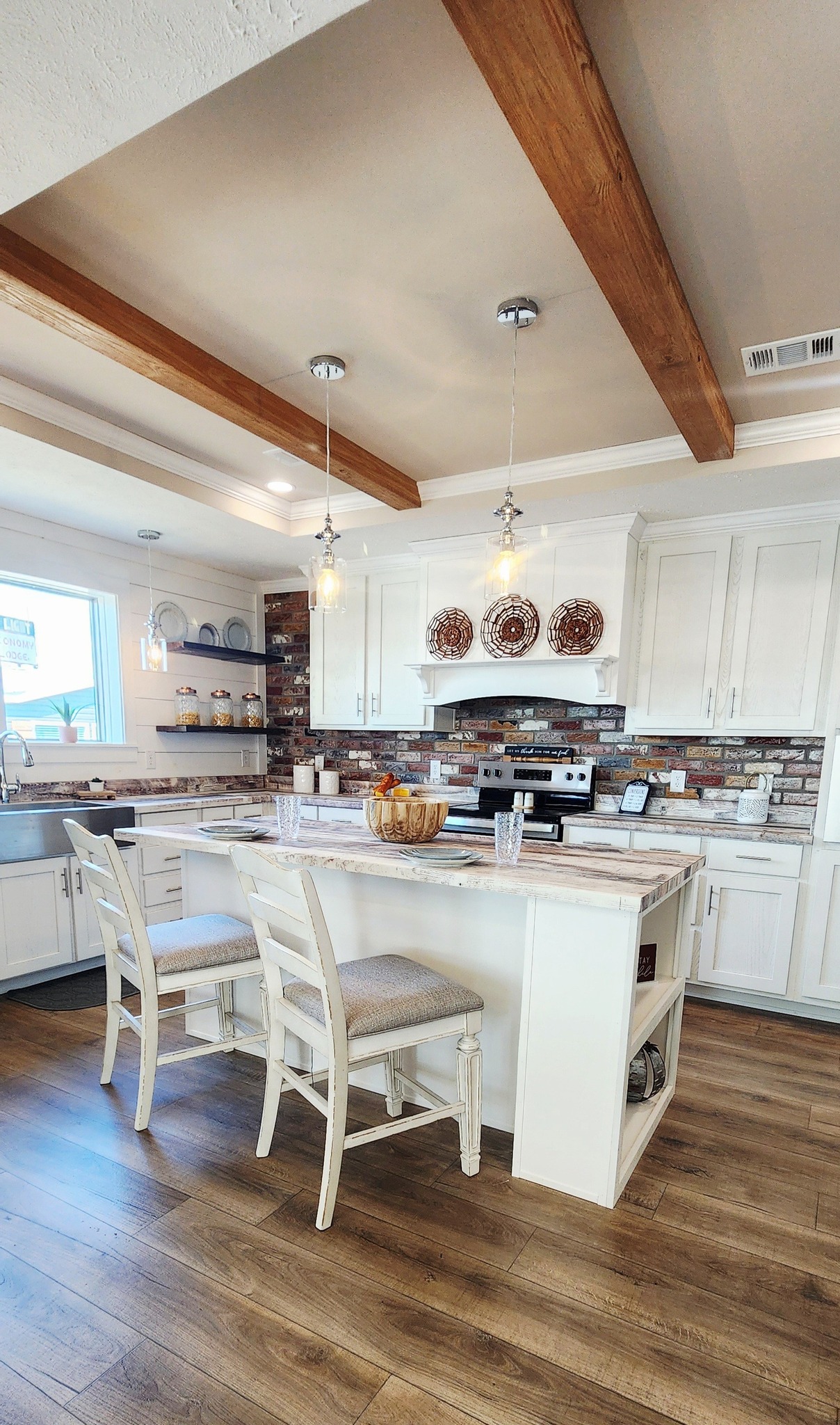 A cozy kitchen with a rustic feel, featuring white cabinets, a brick backsplash, wooden beams, and pendant lights over a central island with two chairs.