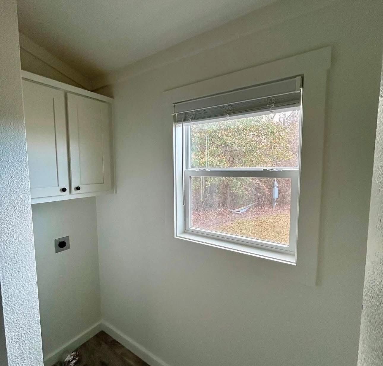 A small, bright laundry room with white walls and cabinets. A window on the right overlooks a yard with trees, casting natural light inside.
