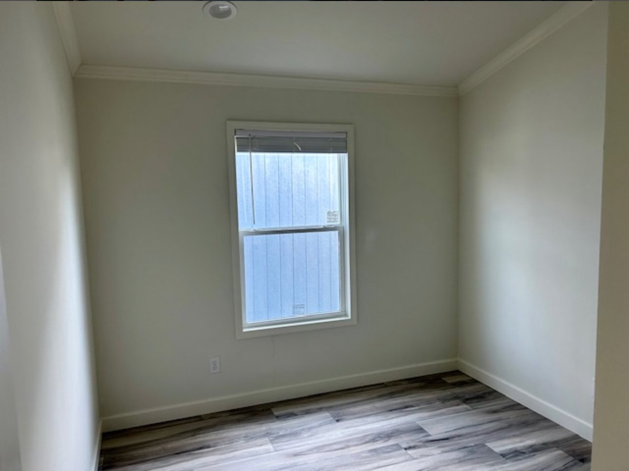 A small, empty room with light gray walls and wood-patterned flooring. A single window with closed blinds lets in minimal light, creating a calm atmosphere.